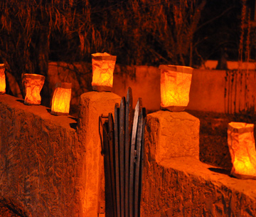 Photo of luminarias glowing atop gate posts on a red clay wall