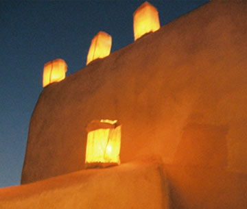 Photo of four golden luminarias glowing dramatically on an adobe wall